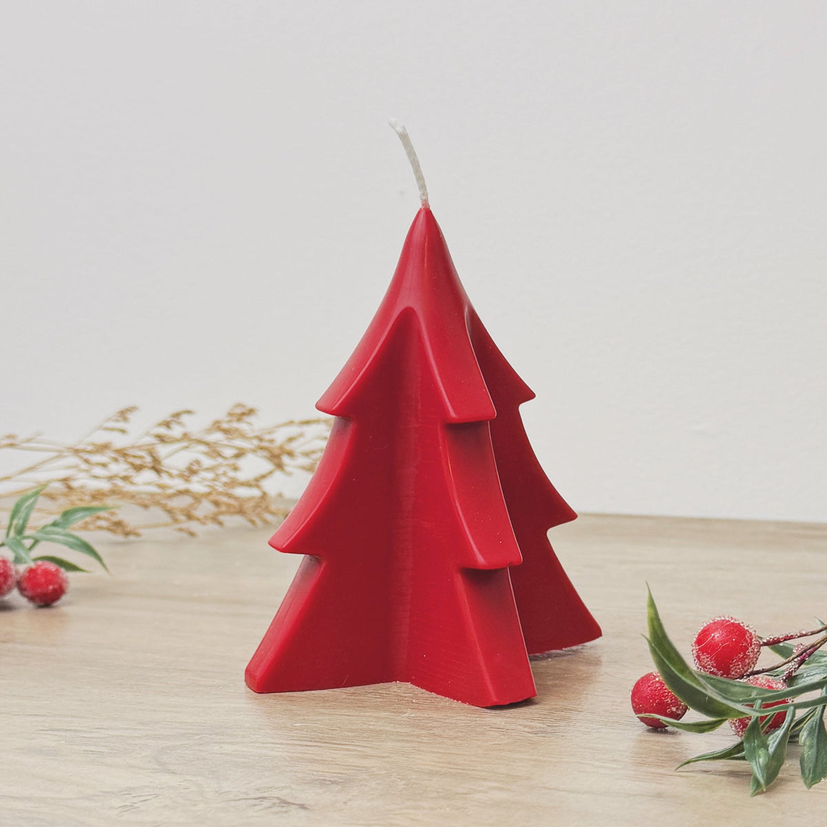 A red Christmas tree-shaped candle with a white wick, sitting on a wooden surface with some festive berries and dried foliage in the background.