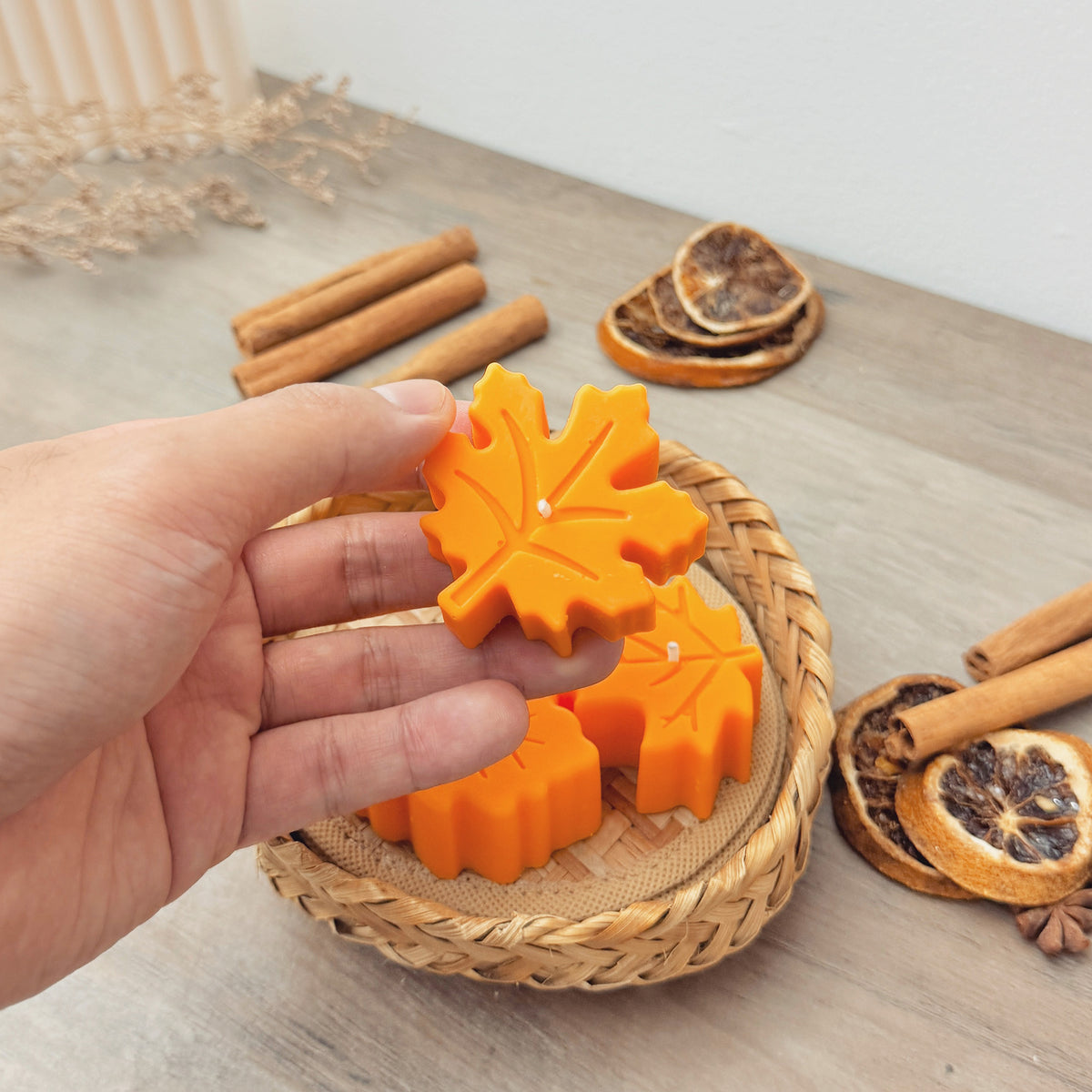 Handmade orange soy tea light candle in a festive maple leaf shape for autumn decor. A hand is holding the candle above a basket, next to cinnamon sticks and dried orange slices on a wooden table.