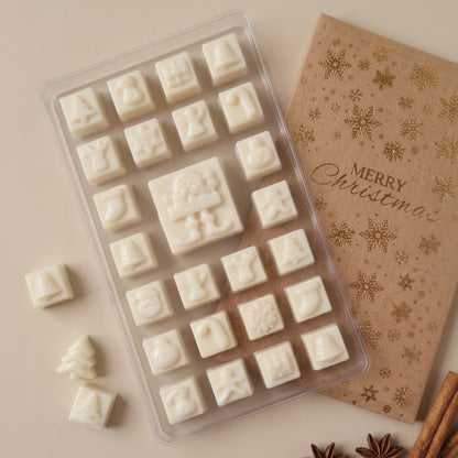 A clear plastic mold filled with small white Christmas-themed soy wax melts shown on a beige background beside a brown kraft card printed with gold snowflakes and the words 'Merry Christmas.'.