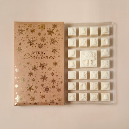 A Christmas advent calender gift box with gold foil snowflakes and the words 'Merry Christmas' next to a clear plastic mold holding white embossed soy wax melt shaped like holiday figures and symbols, on a light beige background.