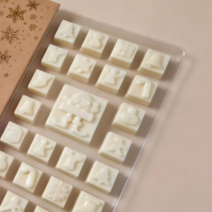 A clear plastic tray holding multiple small white wax melts , each molded with a different holiday-themed embossed design. A brown gift box sleeve with gold snowflakes is partially visible on the left, set against a soft beige background.