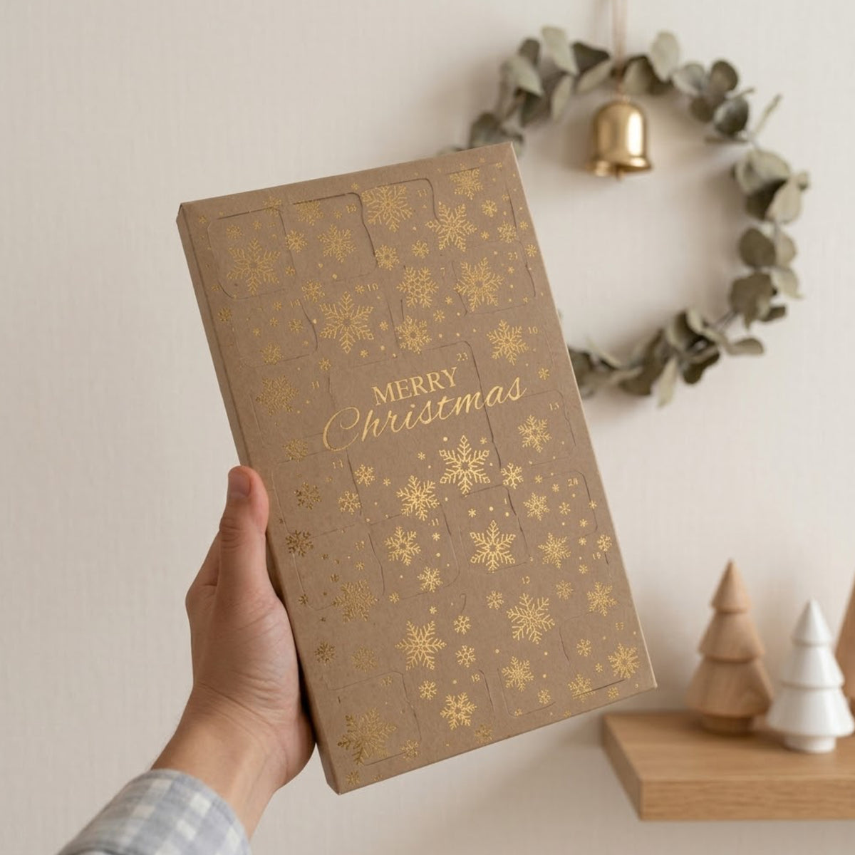 A hand holds a soy wax melt advent calendar decorated with gold foil snowflakes and the words 'MERRY Christmas' in gold script. The background shows a warm, neutral wall with a green eucalyptus wreath and a small wooden shelf with decorative trees.