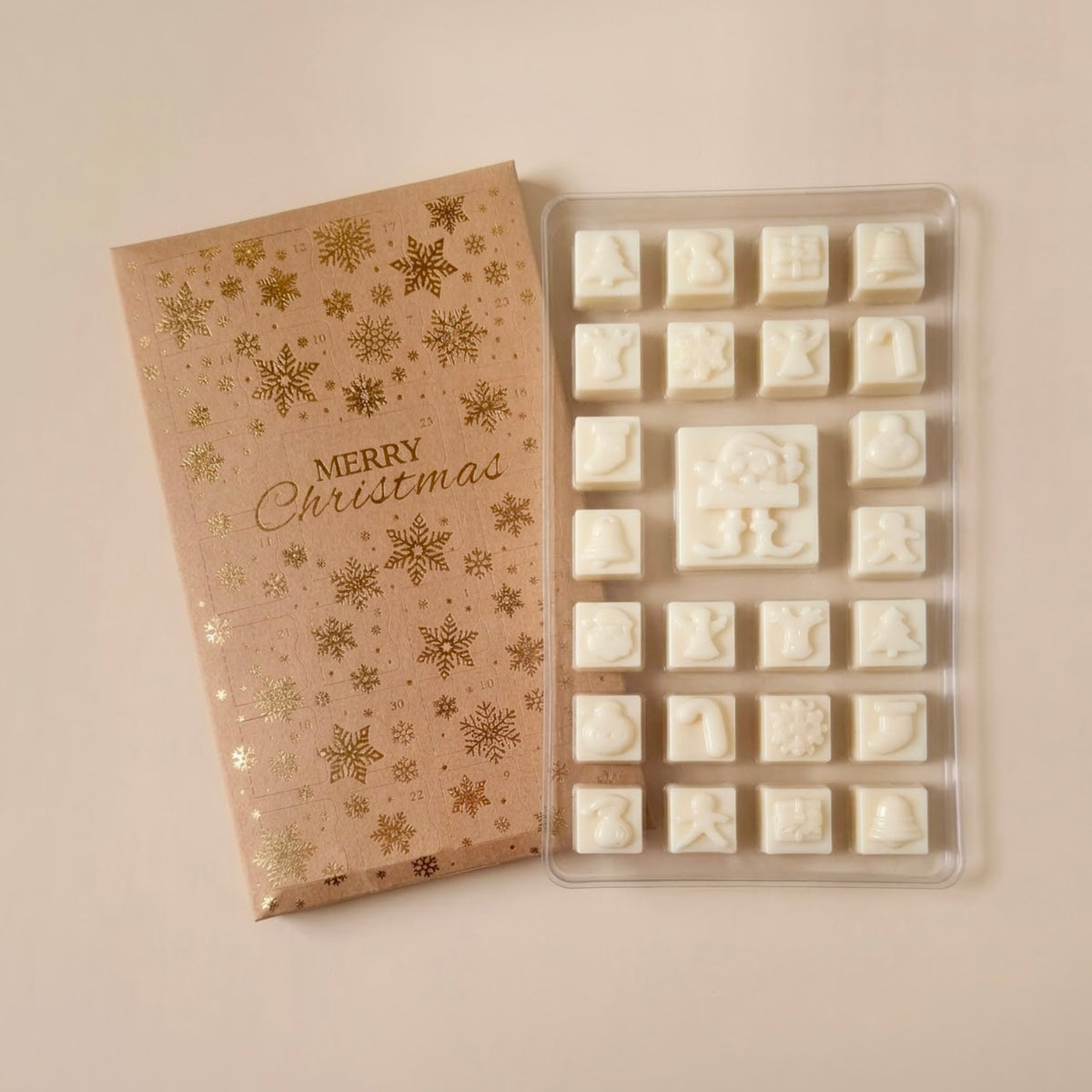 White Christmas-themed soy wax melts arranged in a clear plastic tray next to a gold foil-wrapped gift box with 'Merry Christmas' printed on it, set against a warm beige background.