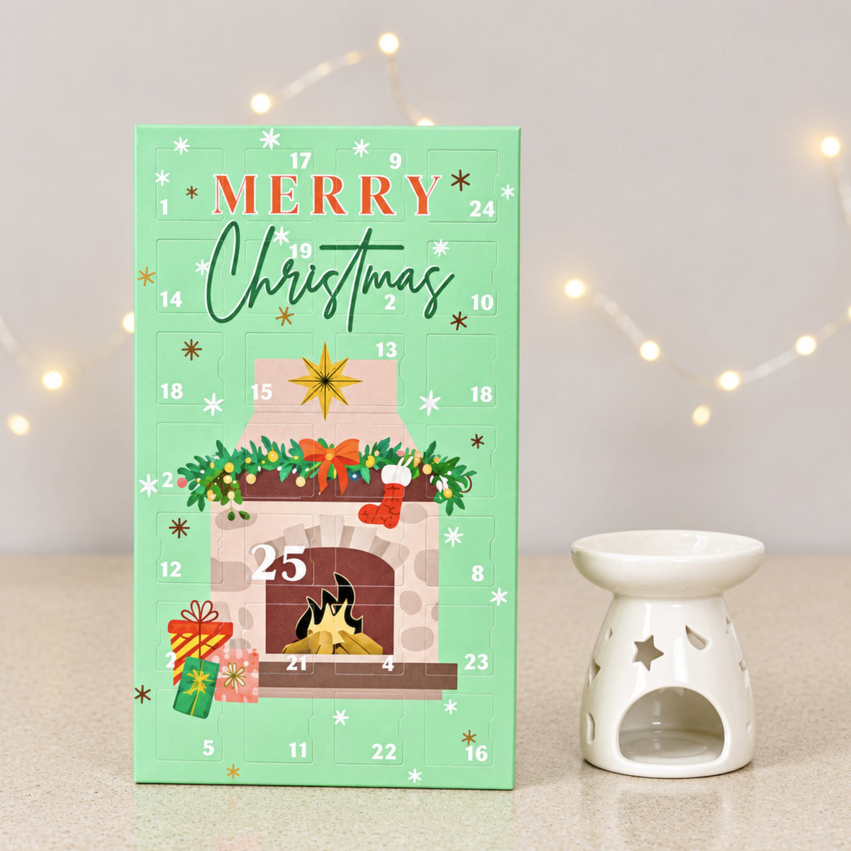 A mint green Christmas wax melt advent calendar with a fireplace illustration and numbered grid, standing on a light surface next to a white ceramic star-shaped candle holder. The background is softly blurred with warm string lights.